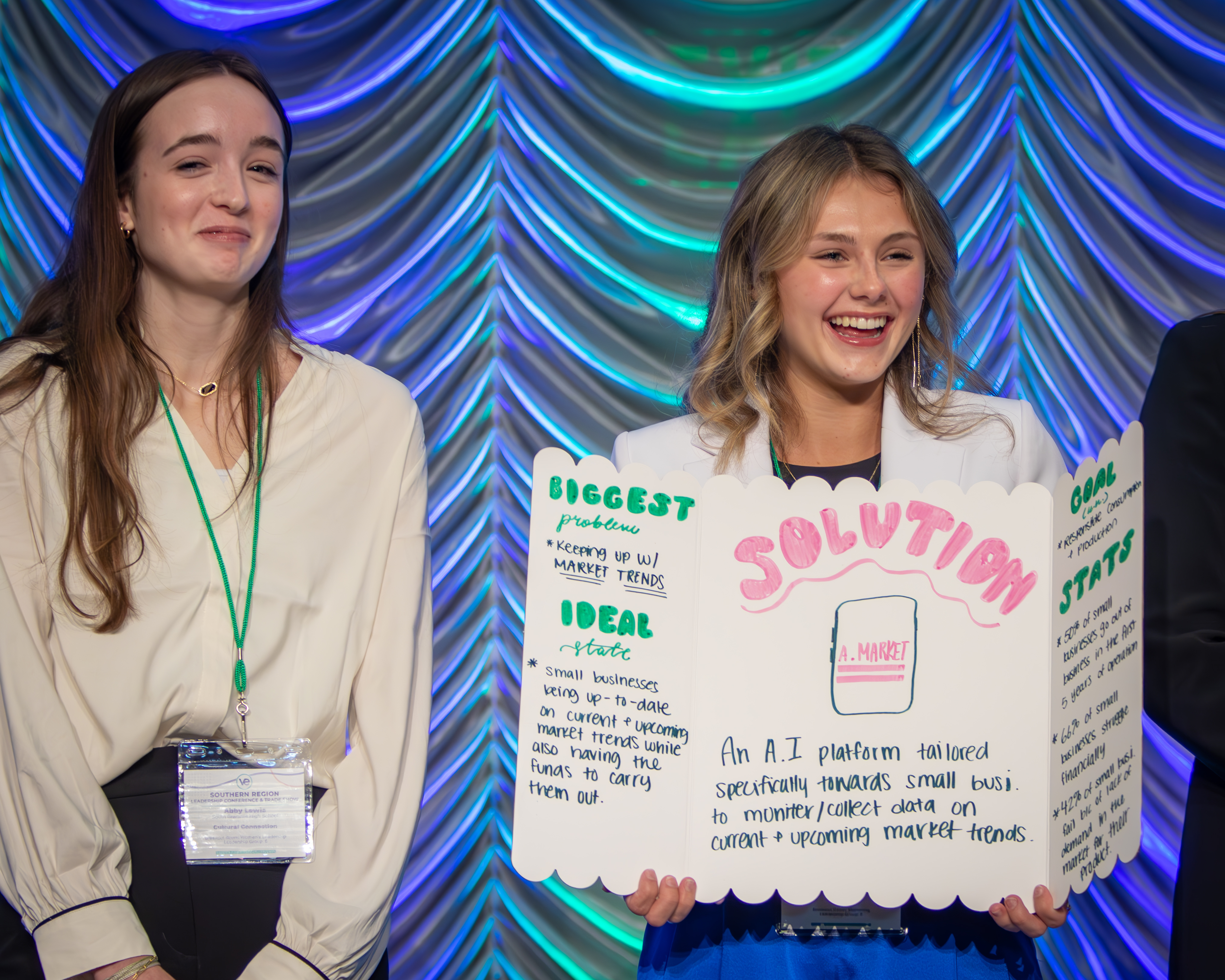 Two female students smile on stage while presenting their business plan. One holds up a poster describing their business.